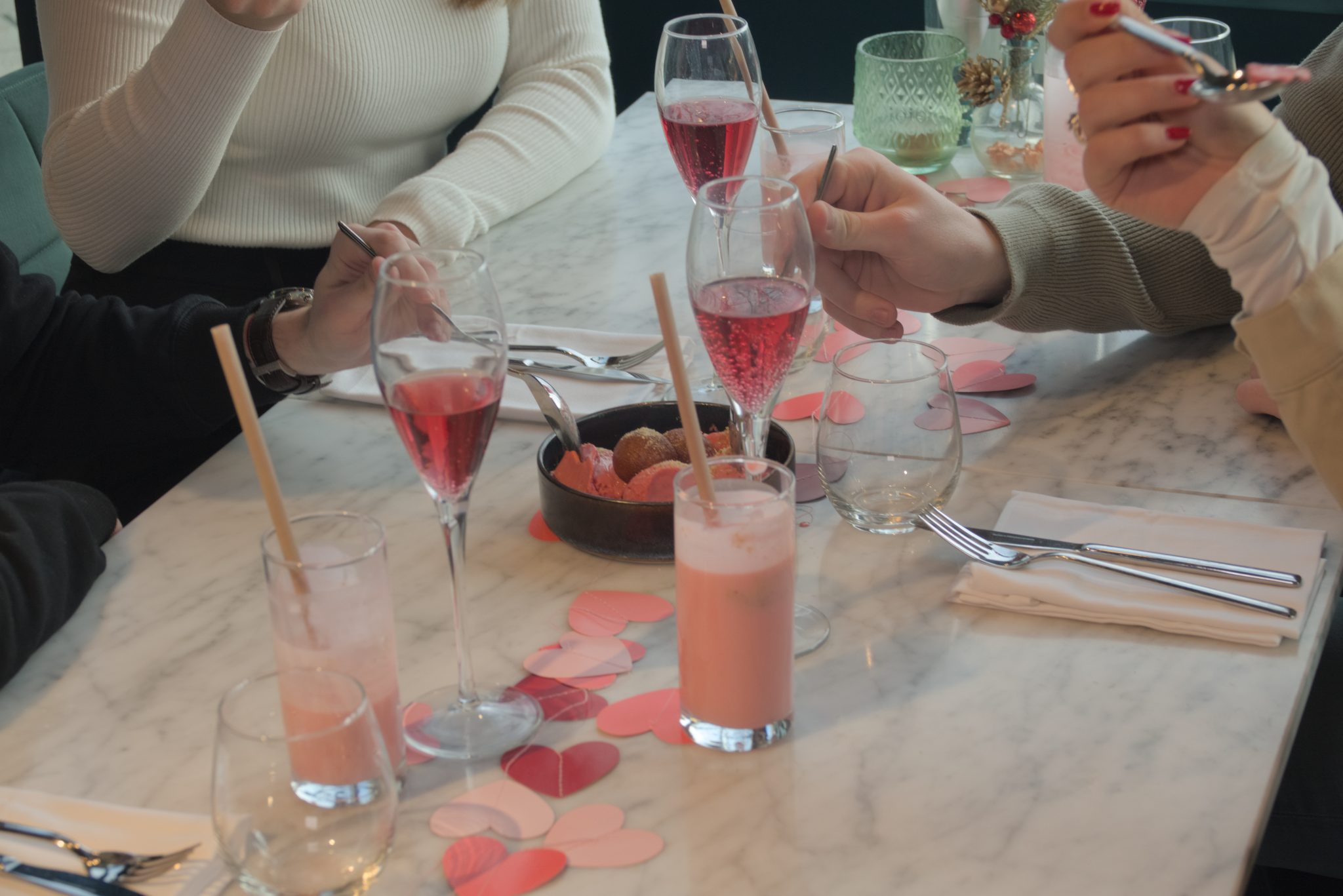 Handen rond een gedeeld dessert, roze drankjes en hartjesdecoratie op een feestelijk gedekte tafel.
