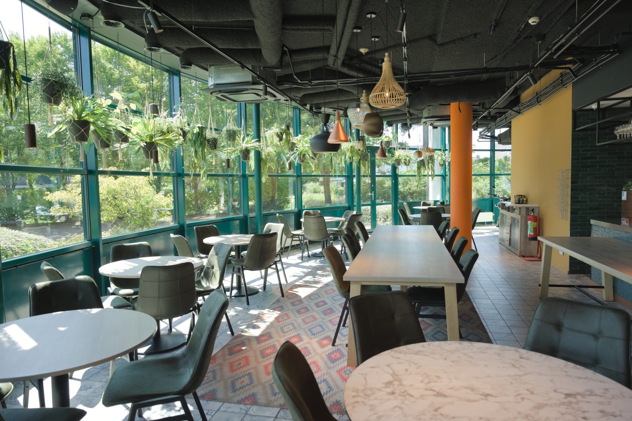 Licht restaurant met grote ramen, hangplanten en ronde en lange tafels.